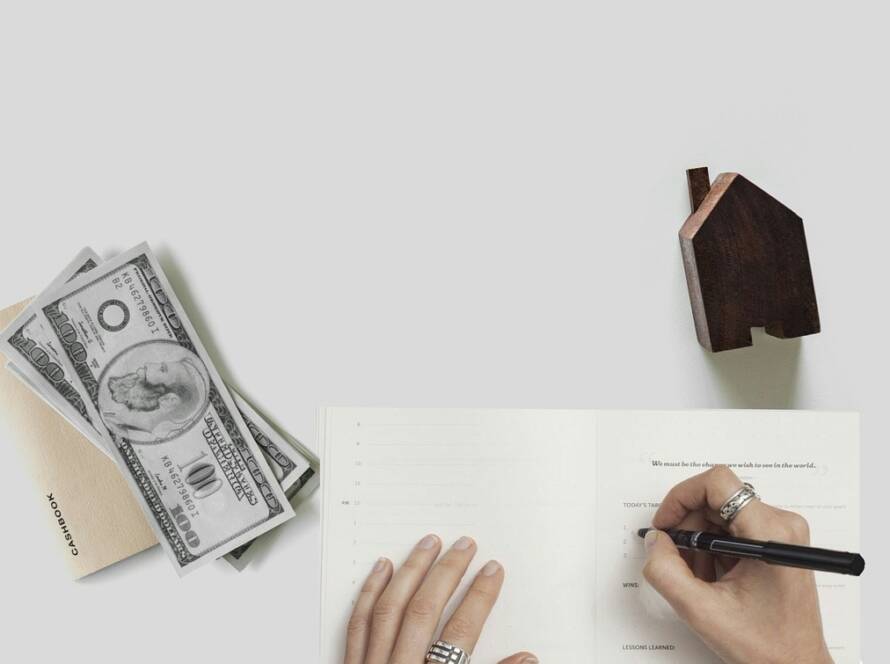 Close-up of hands writing in a notebook with cash, a cashbook, and a wooden house figure representing estate tax liability and real estate disposition in probate.